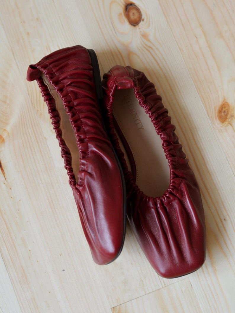 May include: A pair of burgundy leather ballet flats with a gathered, scrunched design. The flats are on a light wood surface.