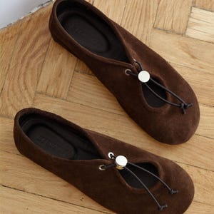 May include: A pair of brown suede ballet flats with a round toe and a drawstring detail. The flats have a small opening on the top of the foot and a silver-colored button. The shoes are on a wooden floor.
