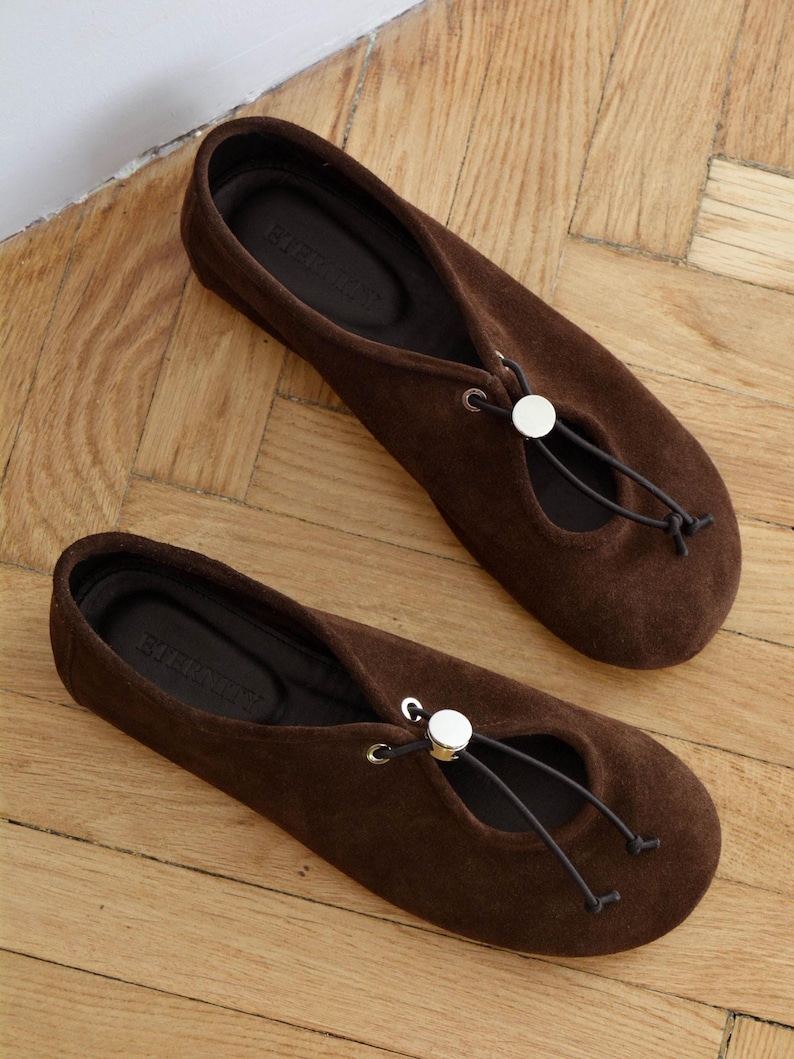 May include: A pair of brown suede ballet flats with a round toe and a drawstring detail. The flats have a small opening on the top of the foot and a silver-colored button. The shoes are on a wooden floor.