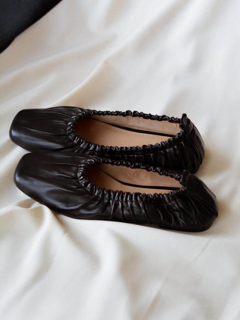 May include: A pair of black leather ballet flats with a gathered, pleated design. The flats have a square toe and a low heel.