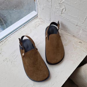 May include: A pair of brown suede slip-on shoes with black straps and buckles. The shoes have a rounded toe and a flat sole.