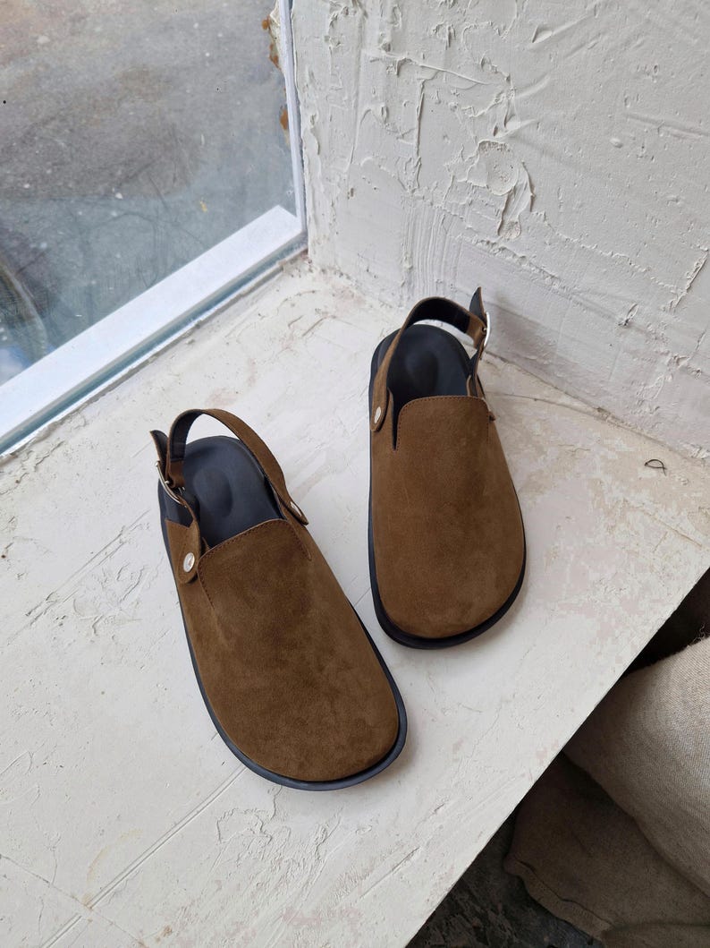 May include: A pair of brown suede slip-on shoes with black straps and buckles. The shoes have a rounded toe and a flat sole.