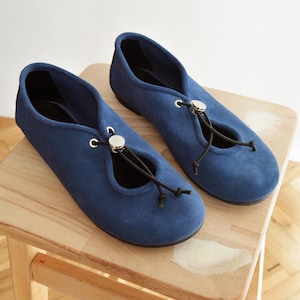 May include: A pair of blue suede ballet flats with a drawstring closure and silver-toned accents. The shoes are displayed on a small wooden stool, showcasing their design and texture. The flats have a classic, minimalist style.