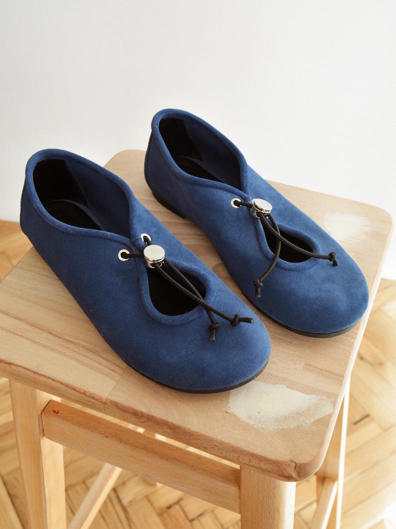 May include: A pair of blue suede ballet flats with a drawstring closure and silver-toned accents. The shoes are displayed on a small wooden stool, showcasing their design and texture. The flats have a classic, minimalist style.