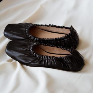 May include: A pair of black leather ballet flats with a gathered, scrunched design. The flats have a square toe and a flat sole.