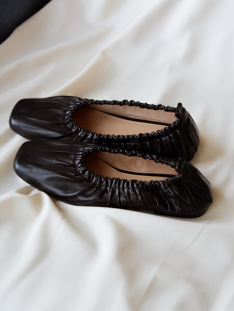 May include: A pair of black leather ballet flats with a gathered, scrunched design. The flats have a square toe and a flat sole.