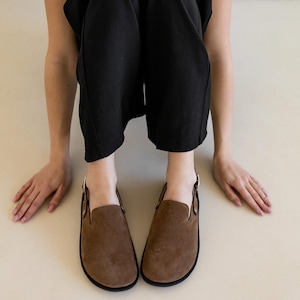May include: A pair of brown suede slip-on shoes with a buckle detail. The shoes have a rounded toe and a flat sole.