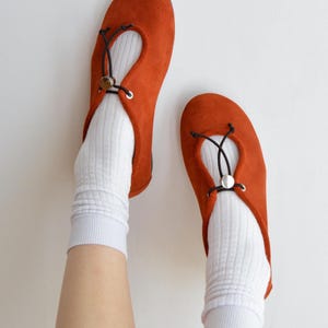 May include: A pair of burnt orange suede ballet flats with a round toe and a black drawstring detail. The shoes are worn with white ribbed socks. The flats have a small silver-colored metal accent.