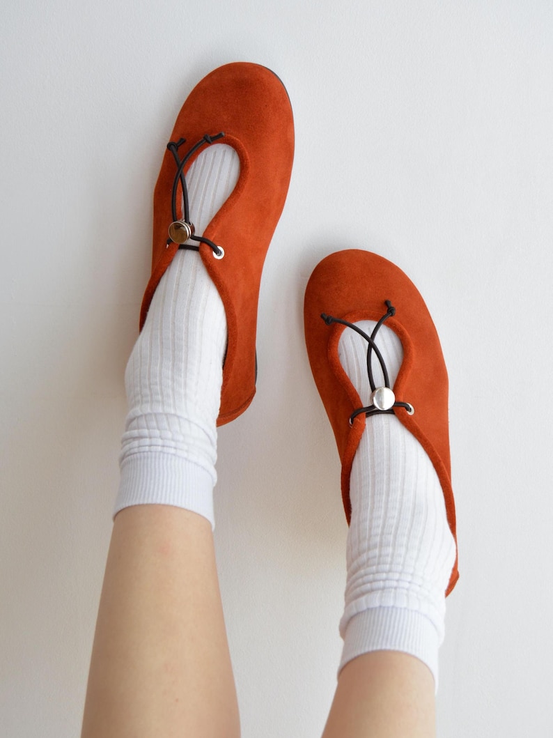 May include: A pair of burnt orange suede ballet flats with a round toe and a black drawstring detail. The shoes are worn with white ribbed socks. The flats have a small silver-colored metal accent.