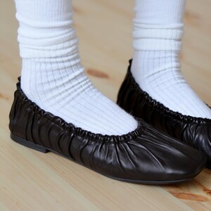 May include: Black leather ballet flats with a gathered detail on the front. The flats are worn with white socks.