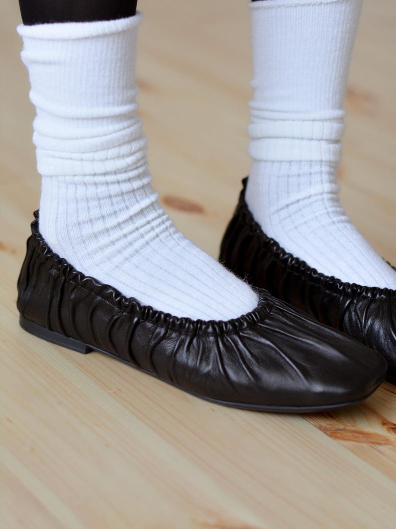 May include: Black leather ballet flats with a gathered detail on the front. The flats are worn with white socks.