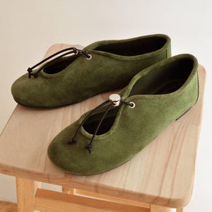 May include: A pair of olive green suede ballet flats with a drawstring closure and a small silver-colored accent. The shoes are displayed on a light wooden stool, showcasing their minimalist design and earthy tones.