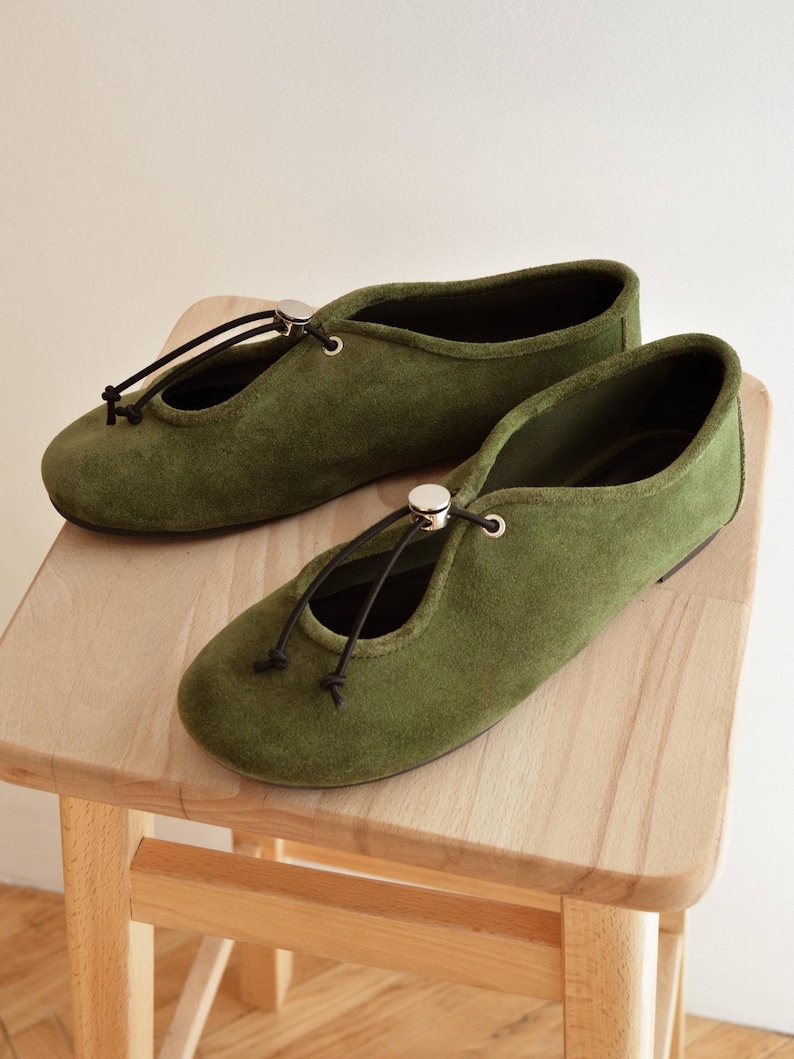 May include: A pair of olive green suede ballet flats with a drawstring closure and a small silver-colored accent. The shoes are displayed on a light wooden stool, showcasing their minimalist design and earthy tones.