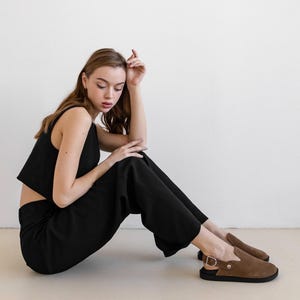 May include: A woman wearing a black crop top and black pants sits on a light beige floor. She is wearing brown suede slip-on shoes with a buckle.