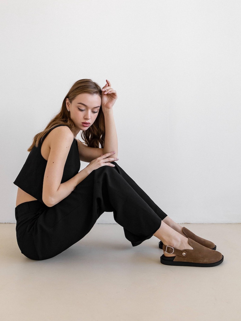 May include: A woman wearing a black crop top and black pants sits on a light beige floor. She is wearing brown suede slip-on shoes with a buckle.