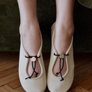 May include: A pair of cream-colored ballet flats with a rounded toe and a decorative black cord and gold button detail. The shoes are made of a soft material and are designed for comfort and style.