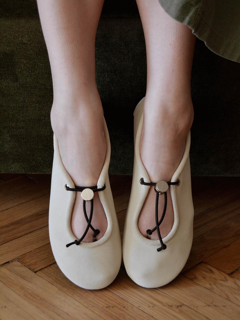 May include: A pair of cream-colored ballet flats with a rounded toe and a decorative black cord and gold button detail. The shoes are made of a soft material and are designed for comfort and style.