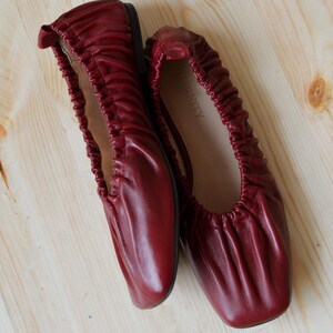 May include: A pair of burgundy leather ballet flats with a gathered, scrunched design. The flats have a square toe and the brand name "INFINITY" is printed on the inside of one shoe.