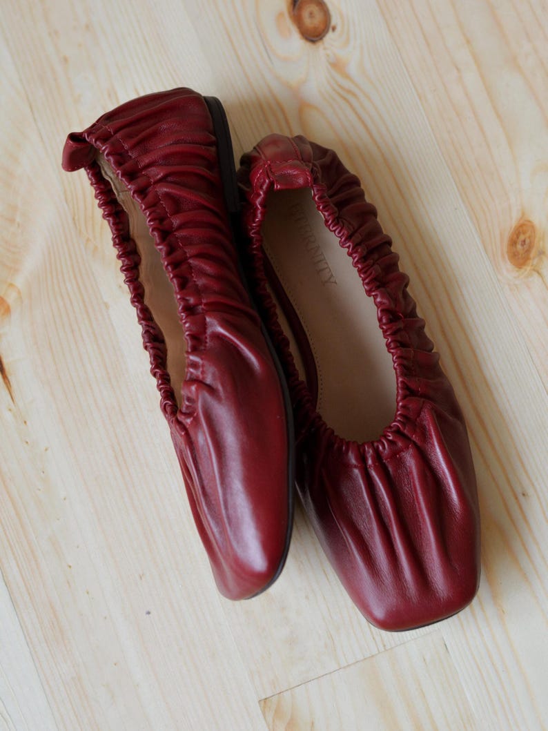 May include: A pair of burgundy leather ballet flats with a gathered, scrunched design. The flats have a square toe and the brand name "INFINITY" is printed on the inside of one shoe.