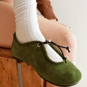 May include: Olive green suede flats with a rounded toe and a black tie closure. The flats are paired with white ribbed socks. The shoes have a small silver ring detail at the top of the foot.