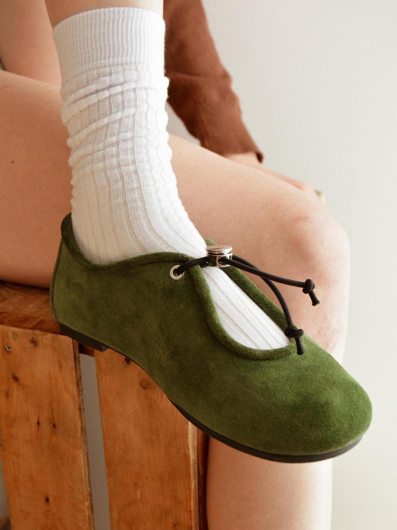 May include: Olive green suede flats with a rounded toe and a black tie closure. The flats are paired with white ribbed socks. The shoes have a small silver ring detail at the top of the foot.