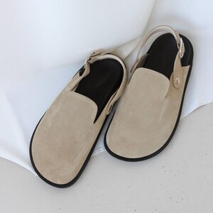 May include: A pair of beige suede slip-on shoes with a slingback strap and black soles.