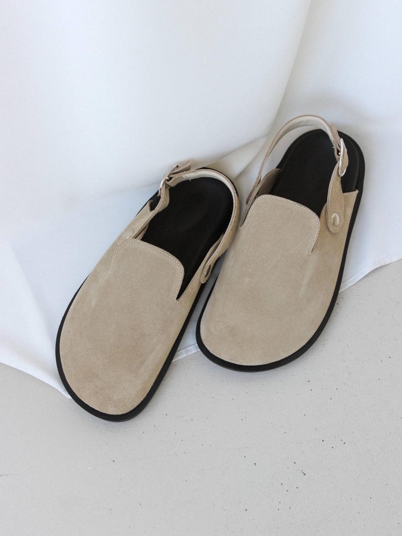 May include: A pair of beige suede slip-on shoes with a slingback strap and black soles.