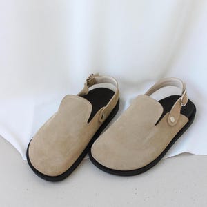 May include: A pair of beige suede slip-on shoes with black soles and a strap around the ankle.