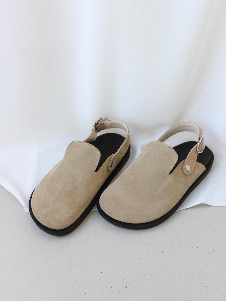 May include: A pair of beige suede slip-on shoes with black soles and a strap around the ankle.