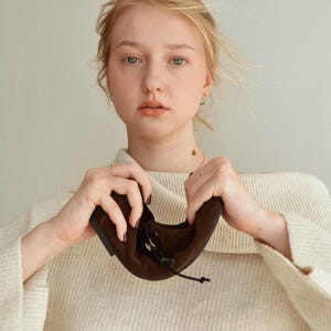 May include: A pair of brown suede flats with a drawstring closure. The shoes are held up by a person wearing a cream-colored sweater. The flats have a rounded toe and a simple design, suitable for everyday wear.
