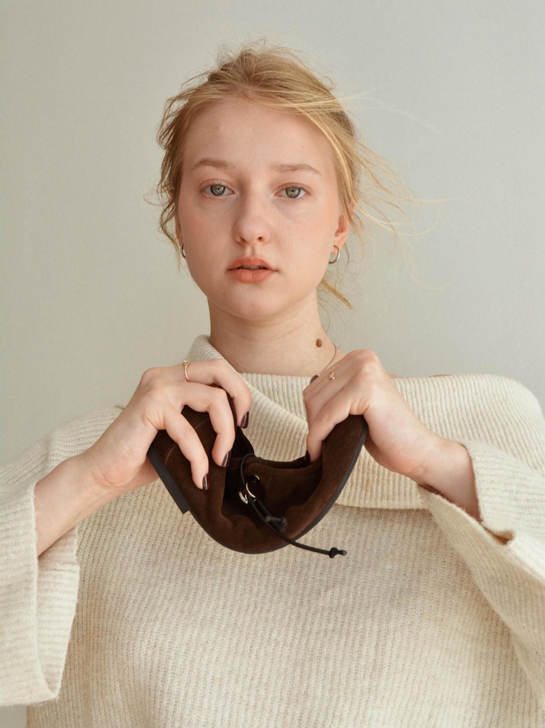 May include: A pair of brown suede flats with a drawstring closure. The shoes are held up by a person wearing a cream-colored sweater. The flats have a rounded toe and a simple design, suitable for everyday wear.