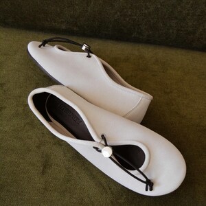 May include: A pair of off-white ballet flats with a black cord detail and a silver-toned button closure. The shoes have a rounded toe and a dark sole. The word "ETERNITY" is printed on the insole.