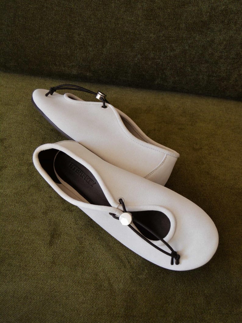 May include: A pair of off-white ballet flats with a black cord detail and a silver-toned button closure. The shoes have a rounded toe and a dark sole. The word "ETERNITY" is printed on the insole.