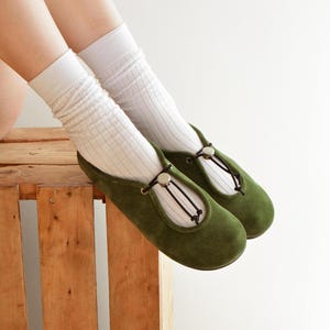 May include: A pair of olive green Mary Jane style shoes with a round toe and a strap closure. The shoes are paired with white ribbed socks. The shoes are resting on a wooden crate.