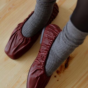 May include: A pair of burgundy leather ballet flats with a gathered elastic top. The flats are worn with gray socks.