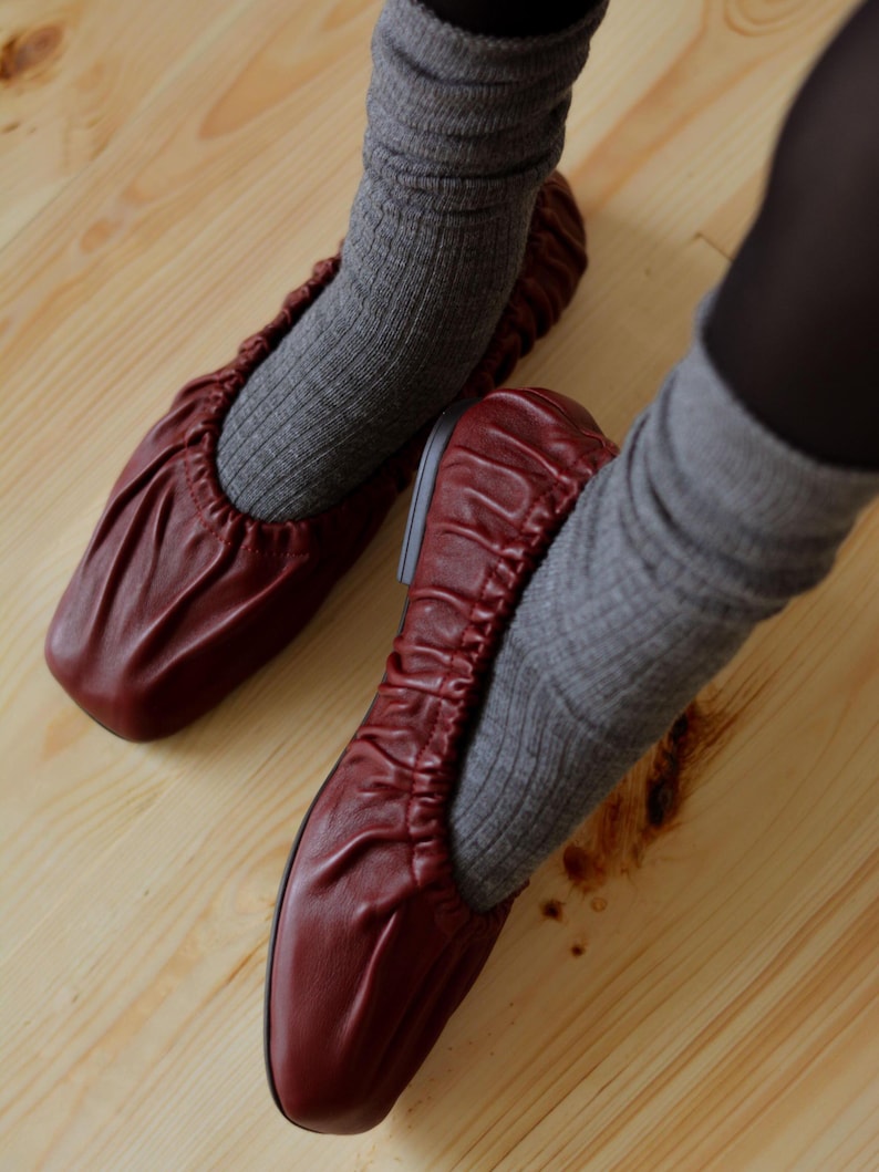 May include: A pair of burgundy leather ballet flats with a gathered elastic top. The flats are worn with gray socks.