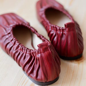 May include: A pair of red leather ballet flats with a gathered detail on the top. The flats are on a wooden surface.