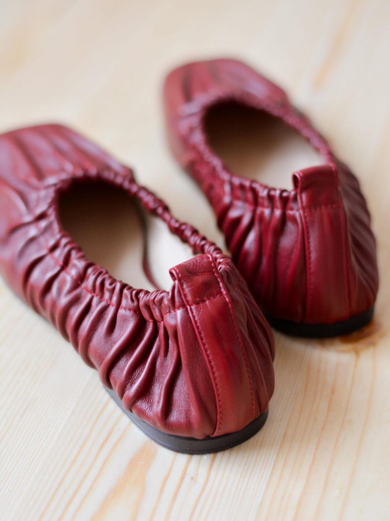 May include: A pair of red leather ballet flats with a gathered detail on the top. The flats are on a wooden surface.