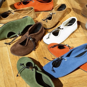 May include: A selection of suede ballet flats in colours such as white, orange, brown, green, and blue. Each flat has a round toe, a small cut-out, and a decorative tie with a metal detail. The flats are displayed on a wooden floor.