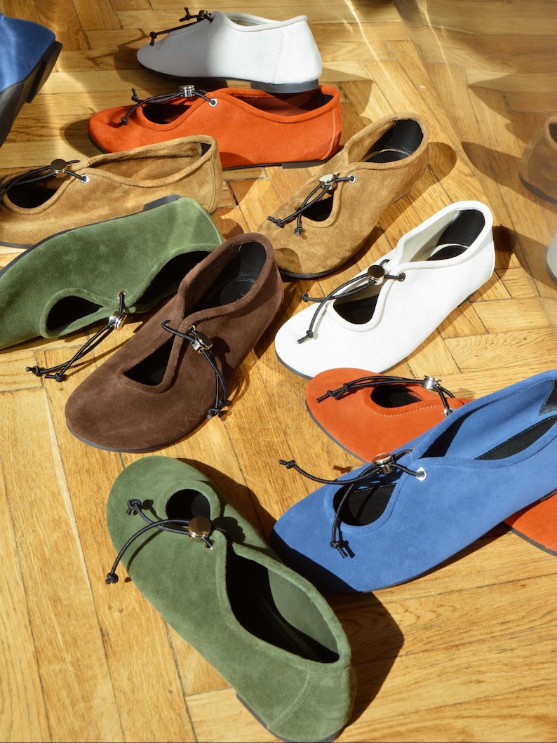 May include: A collection of suede ballet flats in various colors, including white, orange, brown, green, blue, and brown. Each flat features a round toe, a small cut-out, and a decorative tie with a metal accent.