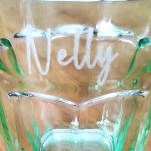 Könnte beinhalten: Ein klares, grün getöntes Glas mit dem Namen "Nelly" in weißer Schrift eingraviert. Das Glas hat ein facettiertes Design und einen dicken Boden. Das Glas ist leer und steht auf einer Holzoberfläche.