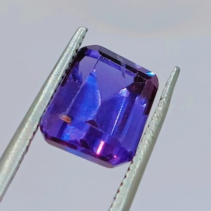 Könnte beinhalten: Ein tiefvioletter Edelstein mit rechteckigem Schliff, gehalten in einer Pinzette. Der Edelstein hat einen leichten Schimmer und ist durchscheinend.