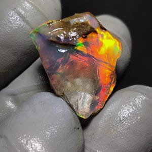 Könnte beinhalten: Ein roher, durchscheinender Opal-Edelstein mit leuchtenden Blitzen in Neon-Orange, Gelb und Grün. Der Stein hat eine unregelmäßige, gezackte Form mit einer teilweise klaren, kristallinen Basis und einem kleinen Fleck aus braunem Muttergestein.