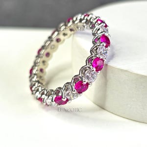 Peut inclure: Une bague en argent ornée de pierres précieuses roses et transparentes alternées. La bague est présentée sur un fond blanc avec le mot "LUXEOTIC" dans le coin supérieur gauche.