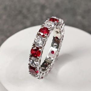 Könnte beinhalten: Ein silberner Eternity-Ring mit abwechselnd roten und klaren Edelsteinen. Der Ring ist mit runden Rubinen und Diamanten besetzt. Der Ring wird auf einer weißen Oberfläche präsentiert. Der Markenname "LUXEOTIC" befindet sich in der oberen linken Ecke.