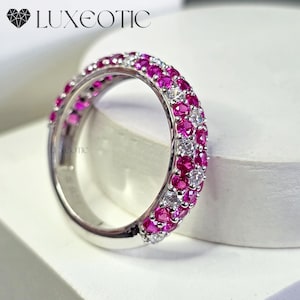 May include: A silver ring with a band of alternating pink gemstones and clear gemstones. The ring is set against a white background. The word "LUXEOTIC" is at the top of the image.