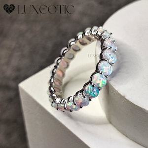May include: A silver band ring with a row of iridescent opal gemstones. The ring is set on a white surface.