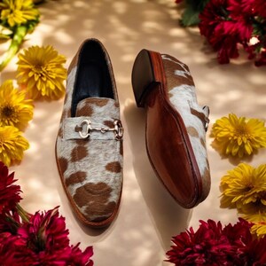 May include: A pair of brown and white cowhide loafers with gold buckles. The loafers are on a light brown background with flowers.