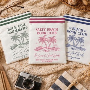 May include: Three white canvas pouches with black zippers and colorful accents. Each pouch features a palm tree design and text: "Book Girl Summer," "Salty Beach Book Club," and "Beach Book Club." The pouches are ideal for carrying books and beach essentials.