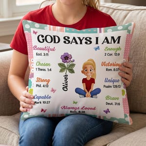 May include: A square throw pillow with a white background featuring the text GOD SAYS I AM, a cartoon illustration of a person with blonde hair, a purple flower, and various inspirational words like Beautiful, Chosen, Strong, and Capable.